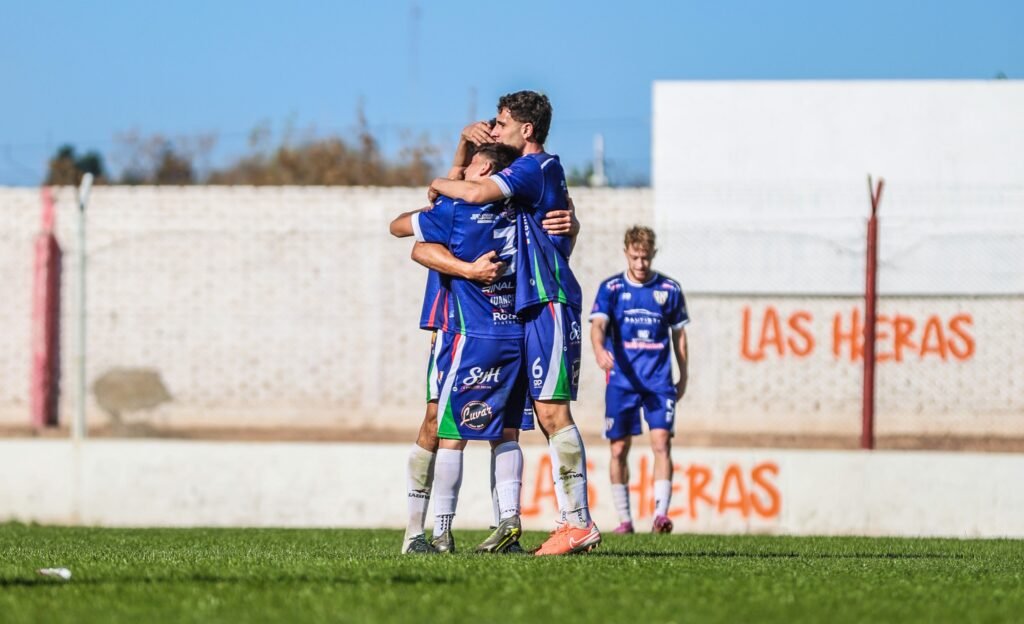 Dos jugadores de Deportivo Guaymallén abrazándose efusivamente para celebrar un gol en la cancha, con un tercer compañero acercándose. De fondo, un muro blanco con la inscripción "Las Heras" en rojo.