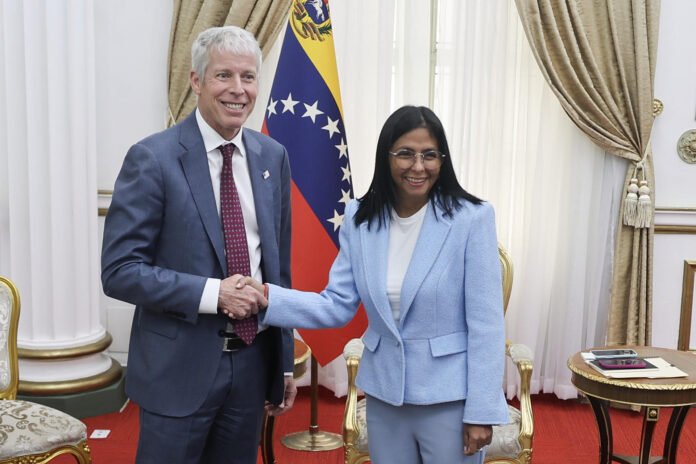 El presidente de EEUU Donald Trump de traje oscuro y corbata roja estrecha la mano de una la presidente de venezuela Delcy Rodriguez con gafas y blazer azul claro frente a la bandera de Venezuela, en un elegante salón durante un encuentro relacionado con el petróleo.  